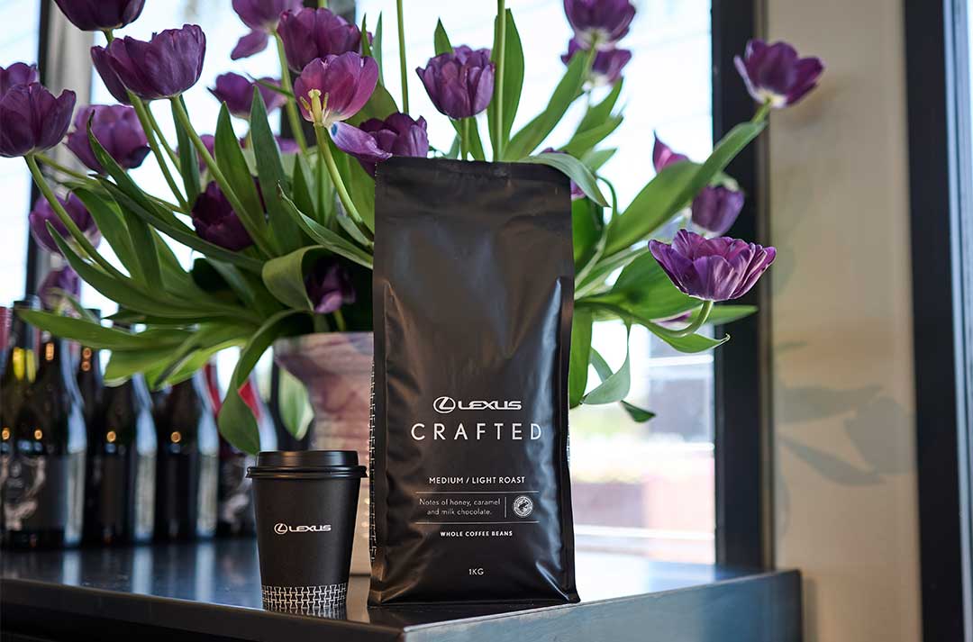 A bag of St Remio x Lexus branded coffee beans on black topped table. A cup of coffee and a white vase holding purple flowers sit alongside.